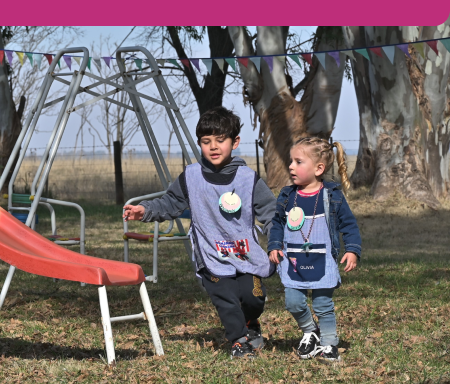 Dos niños pequeños, con delantales escolares y nombres colgados al cuello, caminan juntos en un patio con césped, cerca de un tobogán rojo. Al fondo se ven árboles y una estructura de juego de metal.