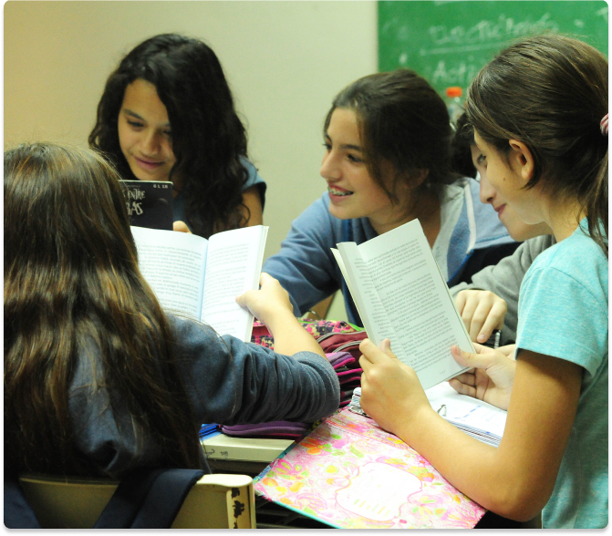 Grupo de estudiantes sentadas alrededor de una mesa en un aula, leyendo y comentando libros, mientras conversan de manera colaborativa.