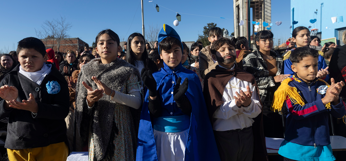 Niños y niñas con trajes tradicionales, entre ellos uno caracterizado como San Martín, aplauden frente a una multitud.